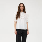 Model i hvid bluse fra Gossia med guldfarvet knap ved kraven, stylet med sorte bukser