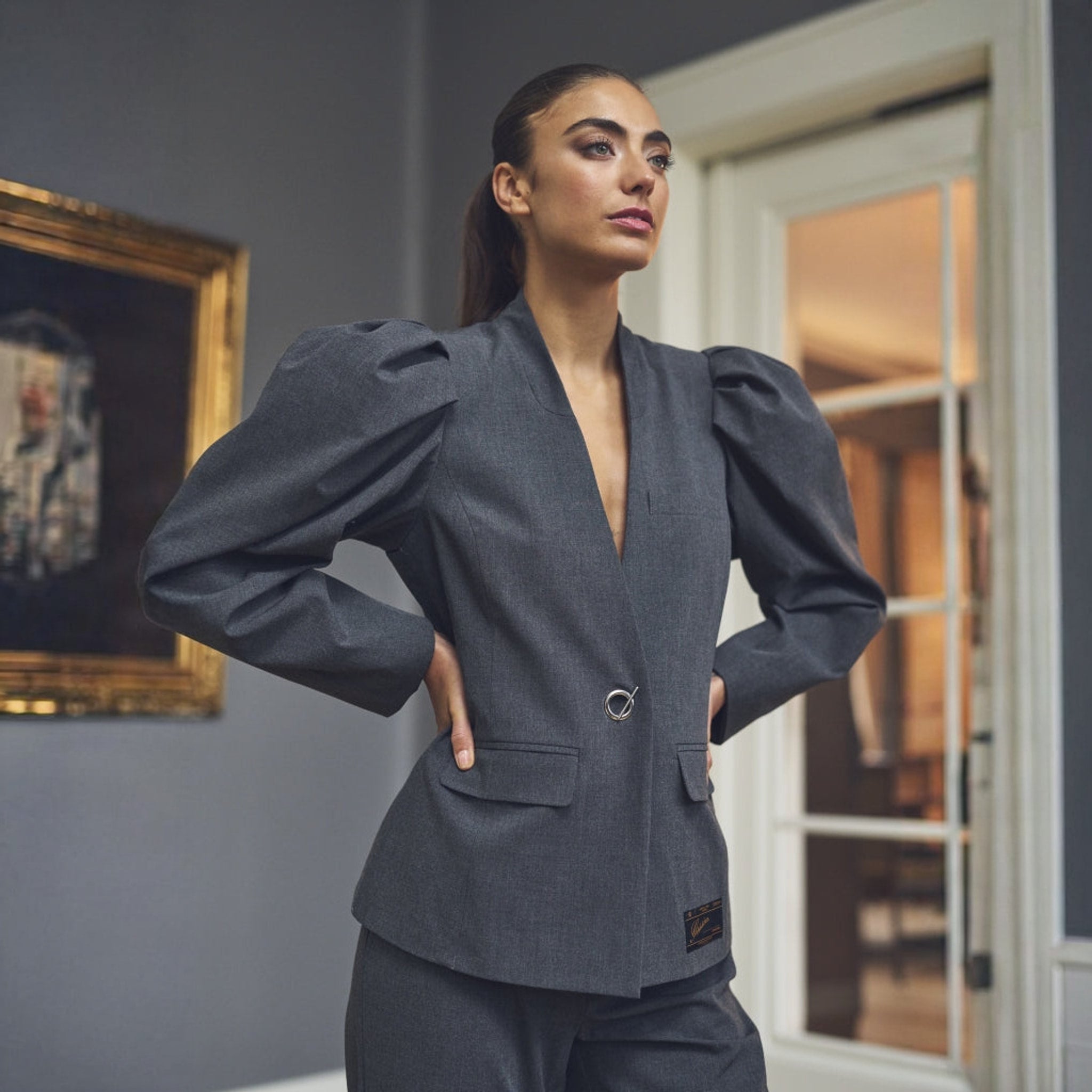Model i grå blazer med puffede ærmer og dyb V-hals, stående med hænderne i siden