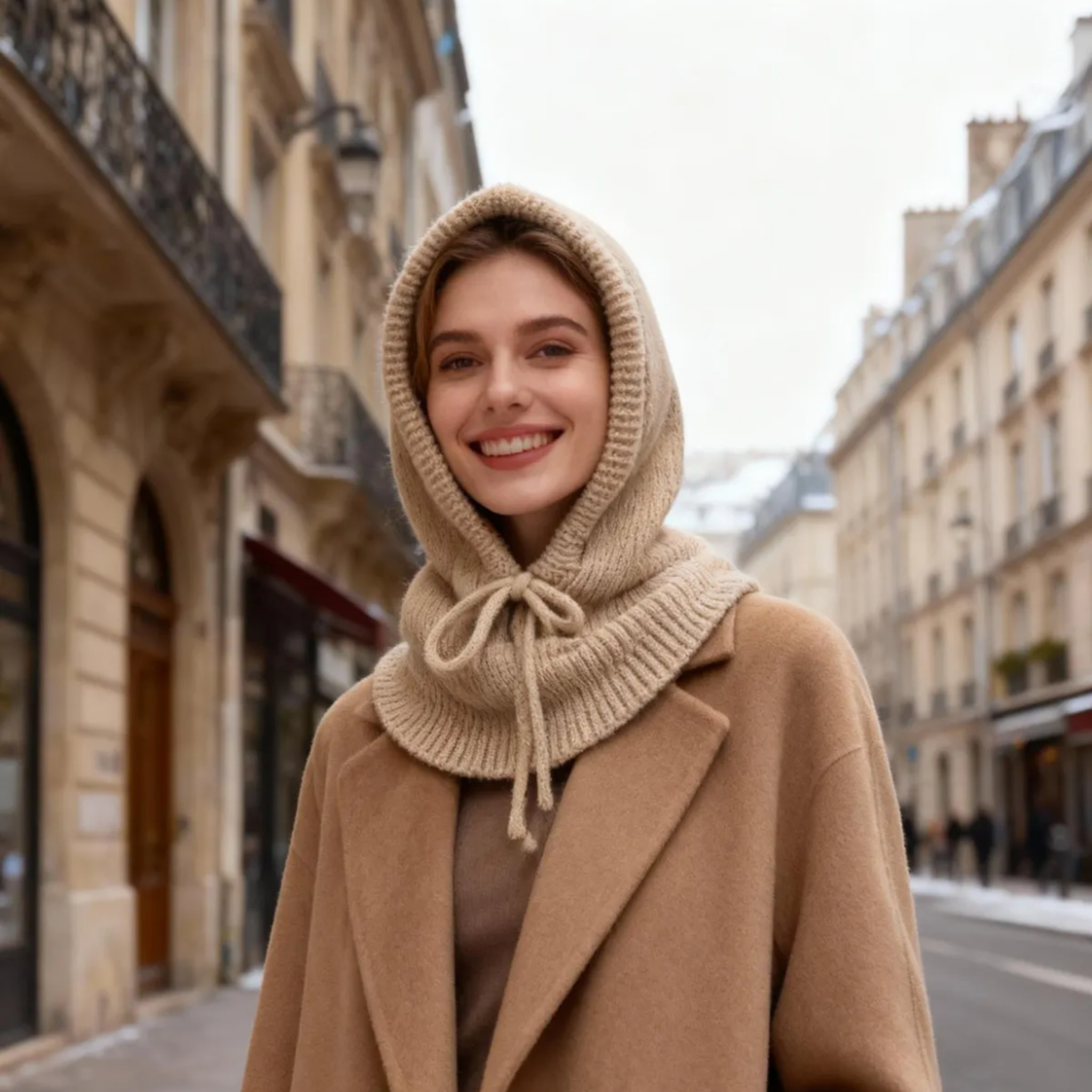 Model med beige balaclava hat i strik med snøre bundet ved halsen, set forfra udendørs