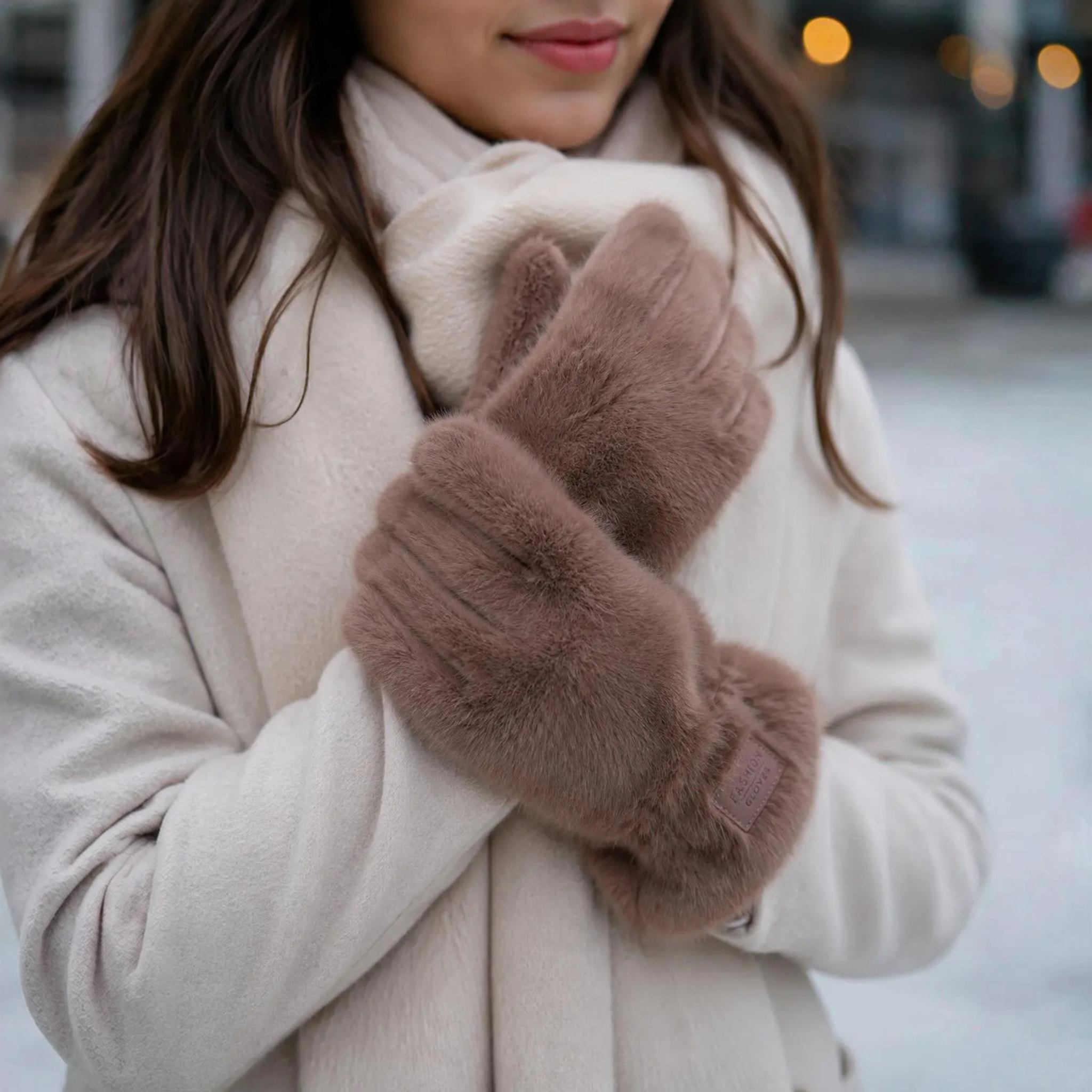 Model med beige handsker i fluffy pelslook i farven Taupe holdt op foran en lys frakke og halstørklæde