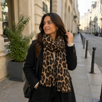 Model med brunt tørklæde med leopardprint viklet om halsen over en sort frakke og en lys strikbluse