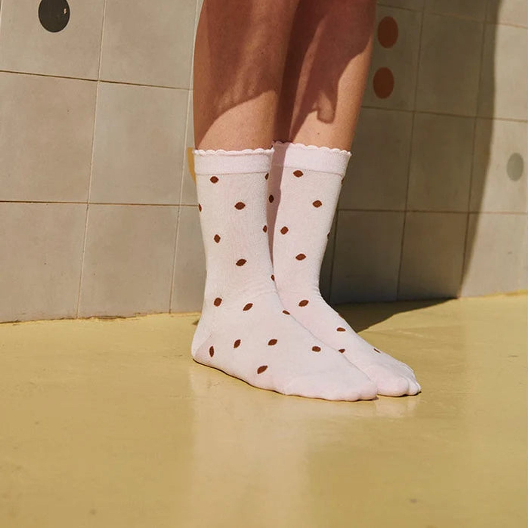 Pink strømper med brune prikker og bølget kant på model uden sko, set forfra ved en flisevæg