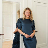Model i navy Benua bluse med puffede ærmer, stylet med satinnederdel, læner sig mod en dør