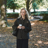 Model i quiltet jakke i khaki og sorte bukser går i en park med kaffe i hånden