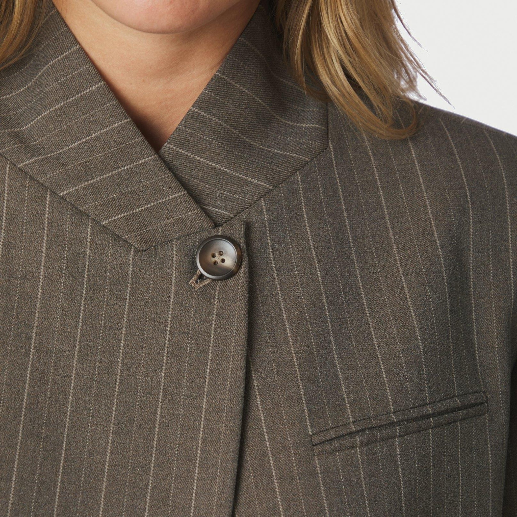 Nærbillede af opstående krave, knap og brystlomme på brun nålestribet blazer