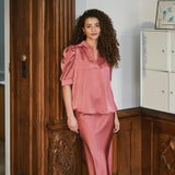 Model i pink satinbluse Roella og lang satinnederdel ved træpanel i en stue