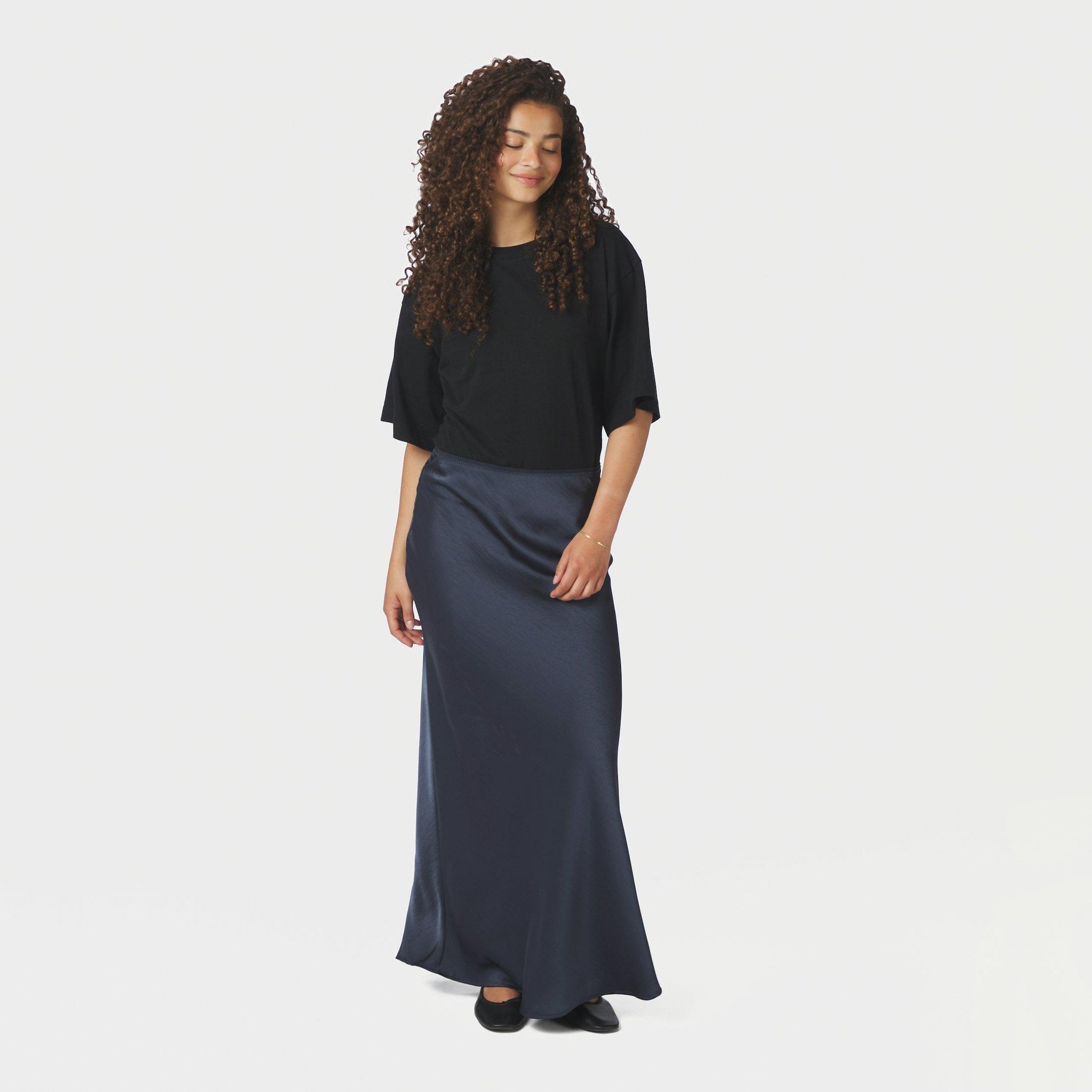 Model i hel figur med lang navy nederdel i satin, sort t-shirt og ballerinaer