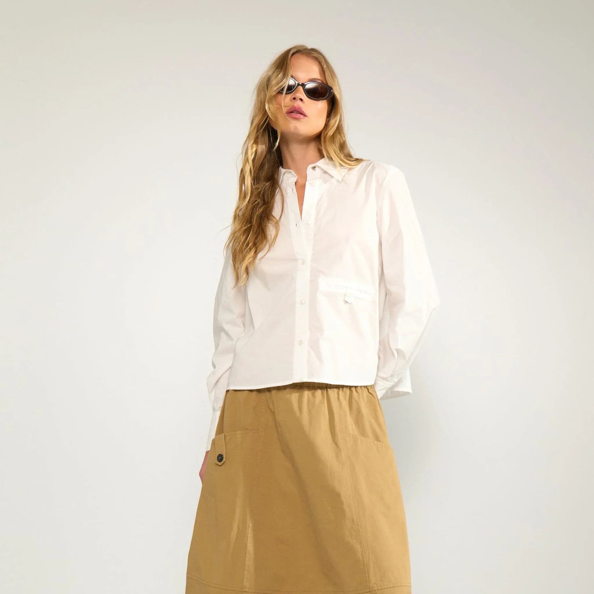 Model i hele figuren i kort hvid skjorte med lange ærmer, stylet med beige midi-nederdel
