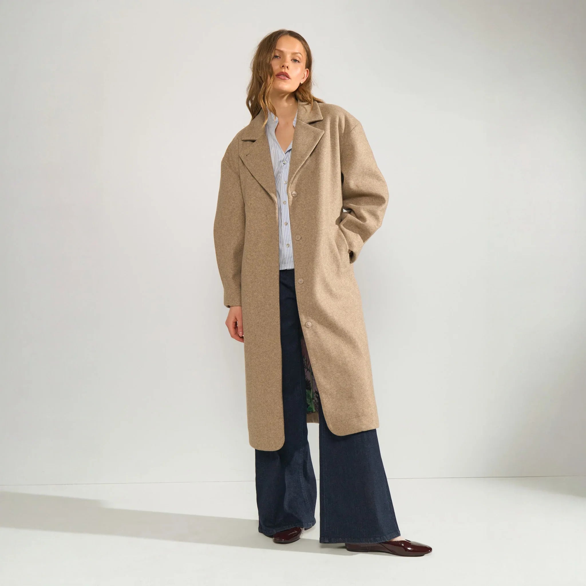 Model i åben lang beige jakke med stribet skjorte og wide leg jeans, set forfra