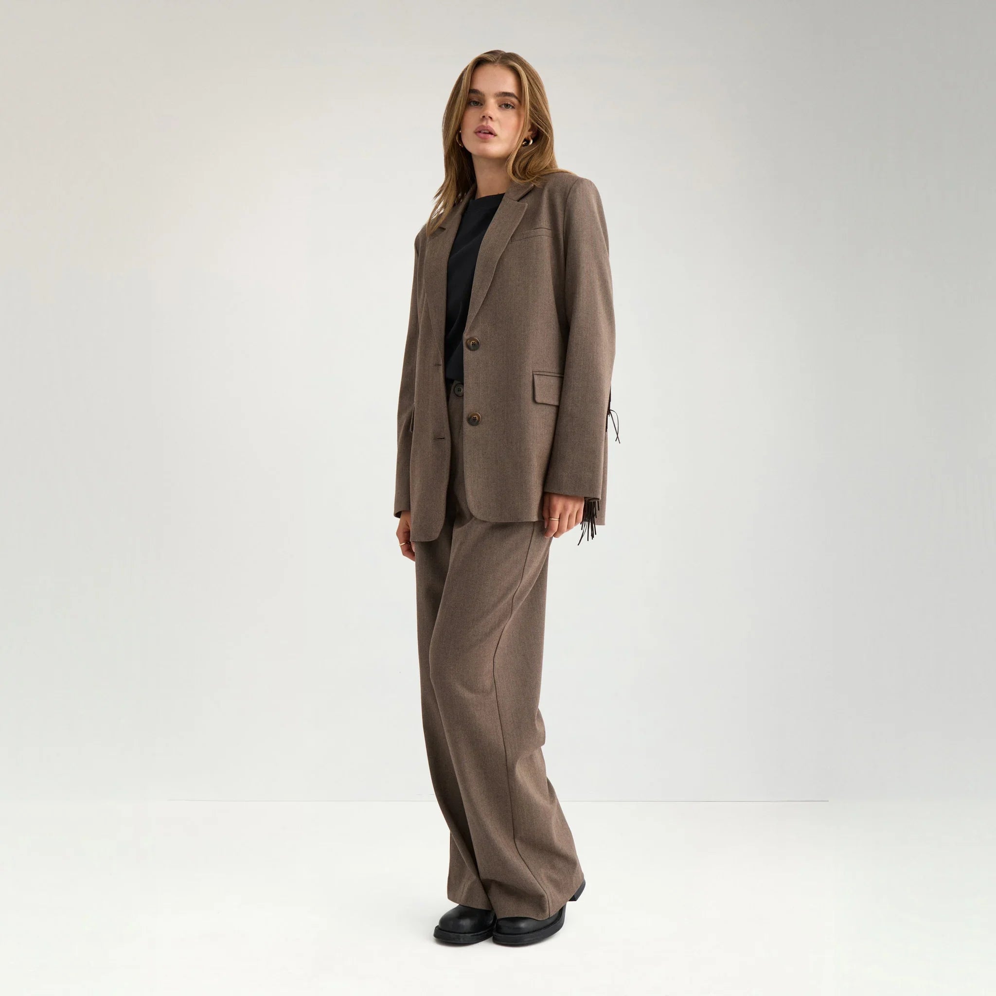 Model i brun blazer med frynser, sort t-shirt og brune bukser, hele figuren