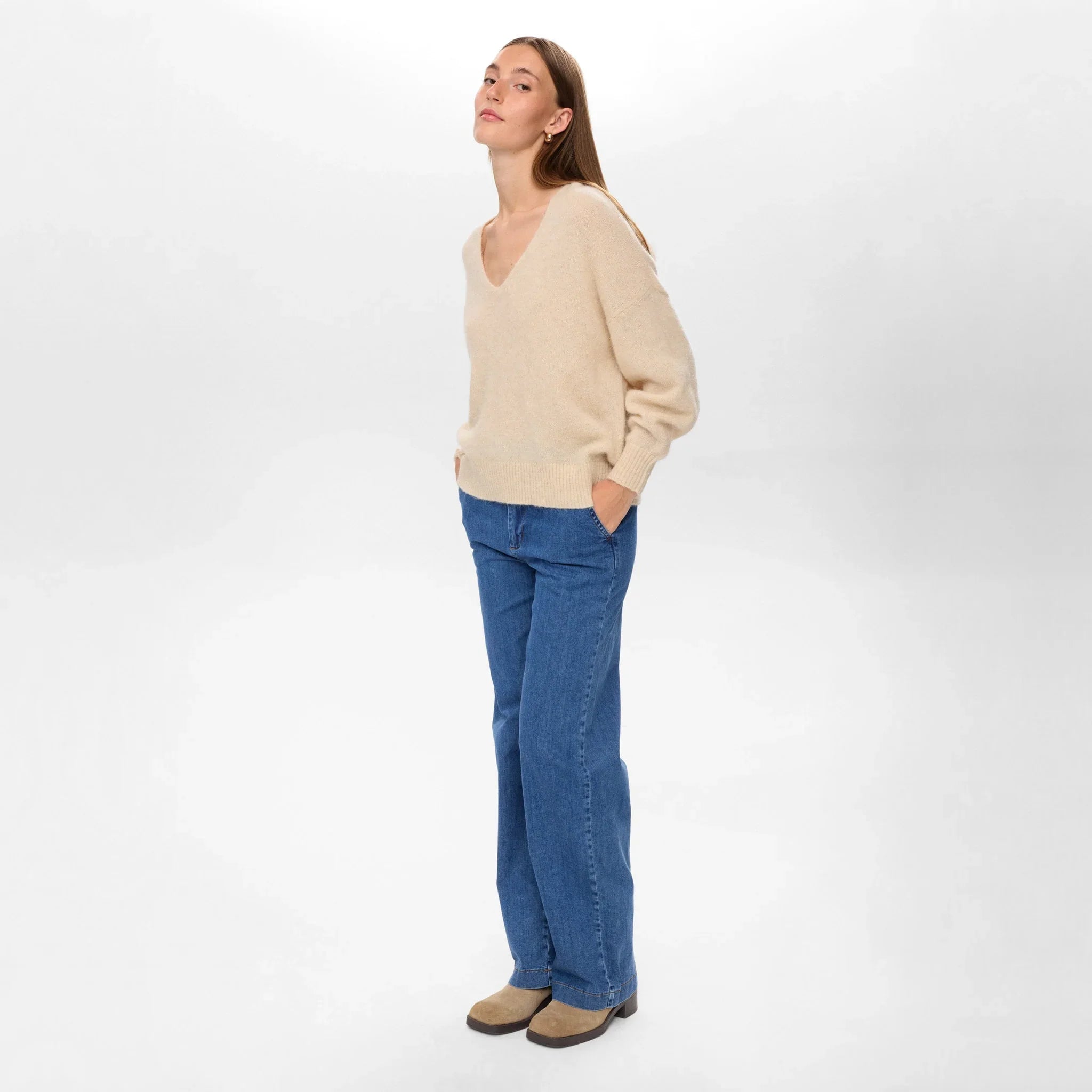 Model i beige pullover med V-hals set fra siden i fuld længde med jeans