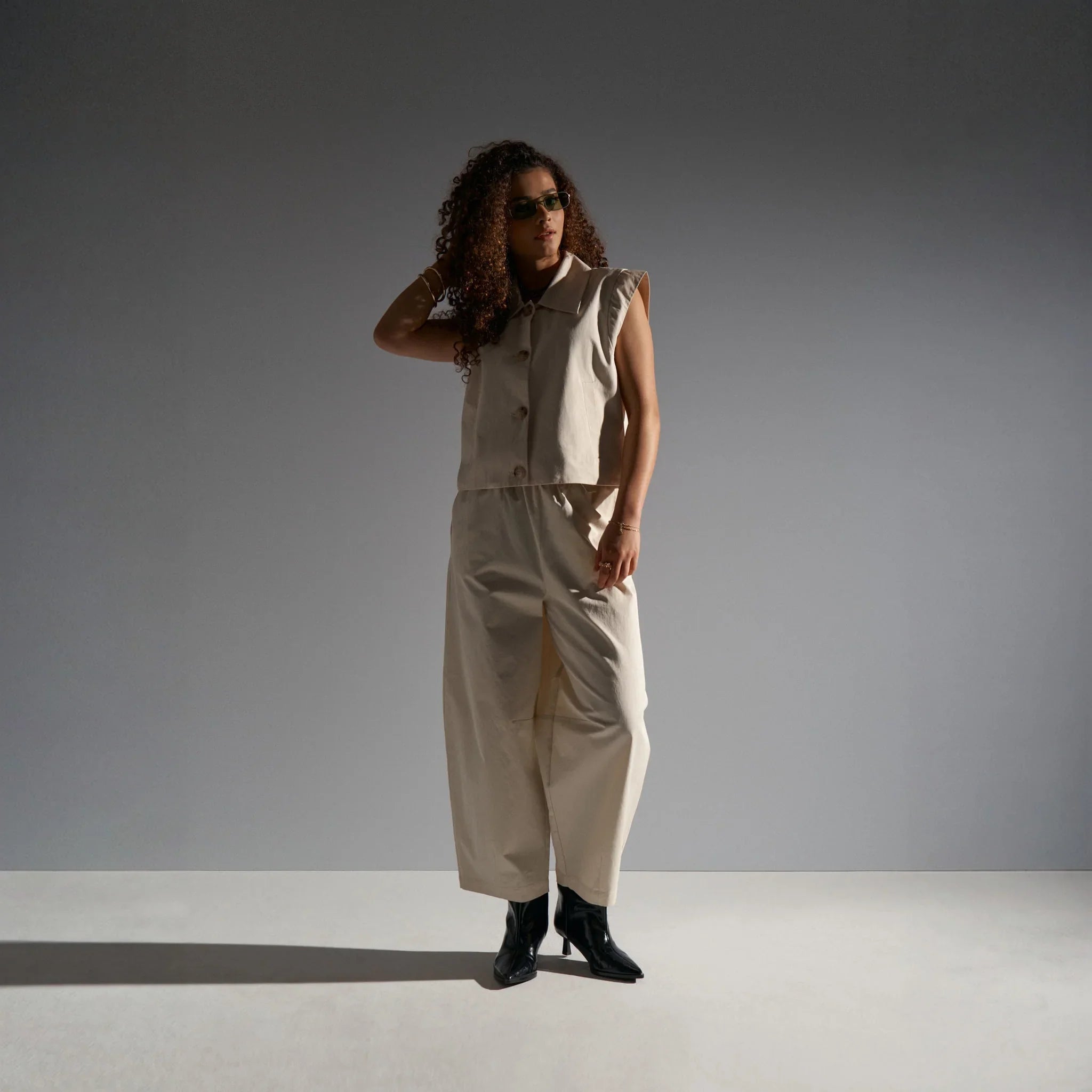 Model i beige wide leg bukser med buet form mod grå baggrund