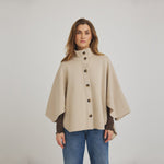 Model i beige poncho-inspireret strik med knapper foran, stylet med brun langærmet top og blå jeans