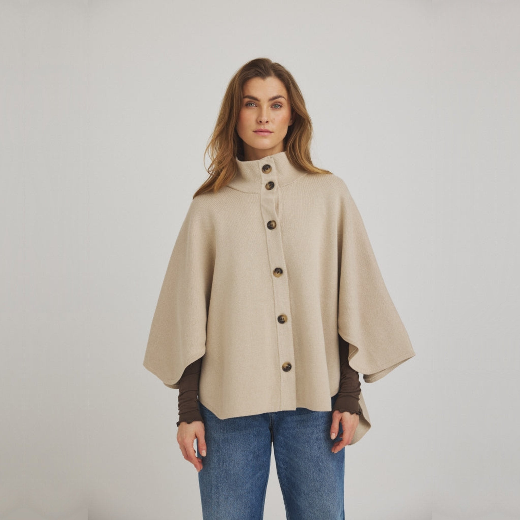 Model i beige poncho-inspireret strik med knapper foran, stylet med brun langærmet top og blå jeans