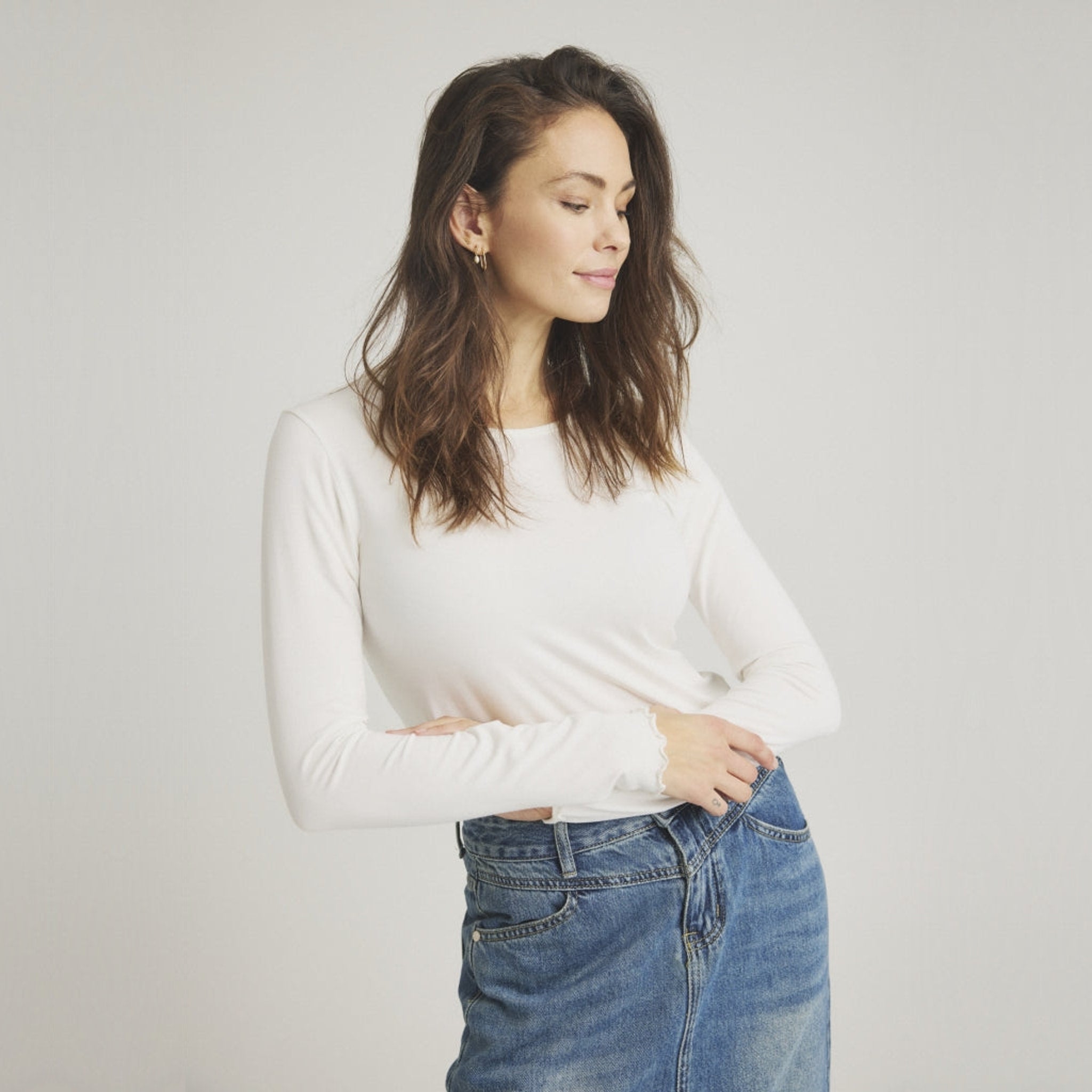 Model i tætsiddende hvid langærmet t-shirt med bølgede kanter ved ærmerne, stylet med denimnederdel