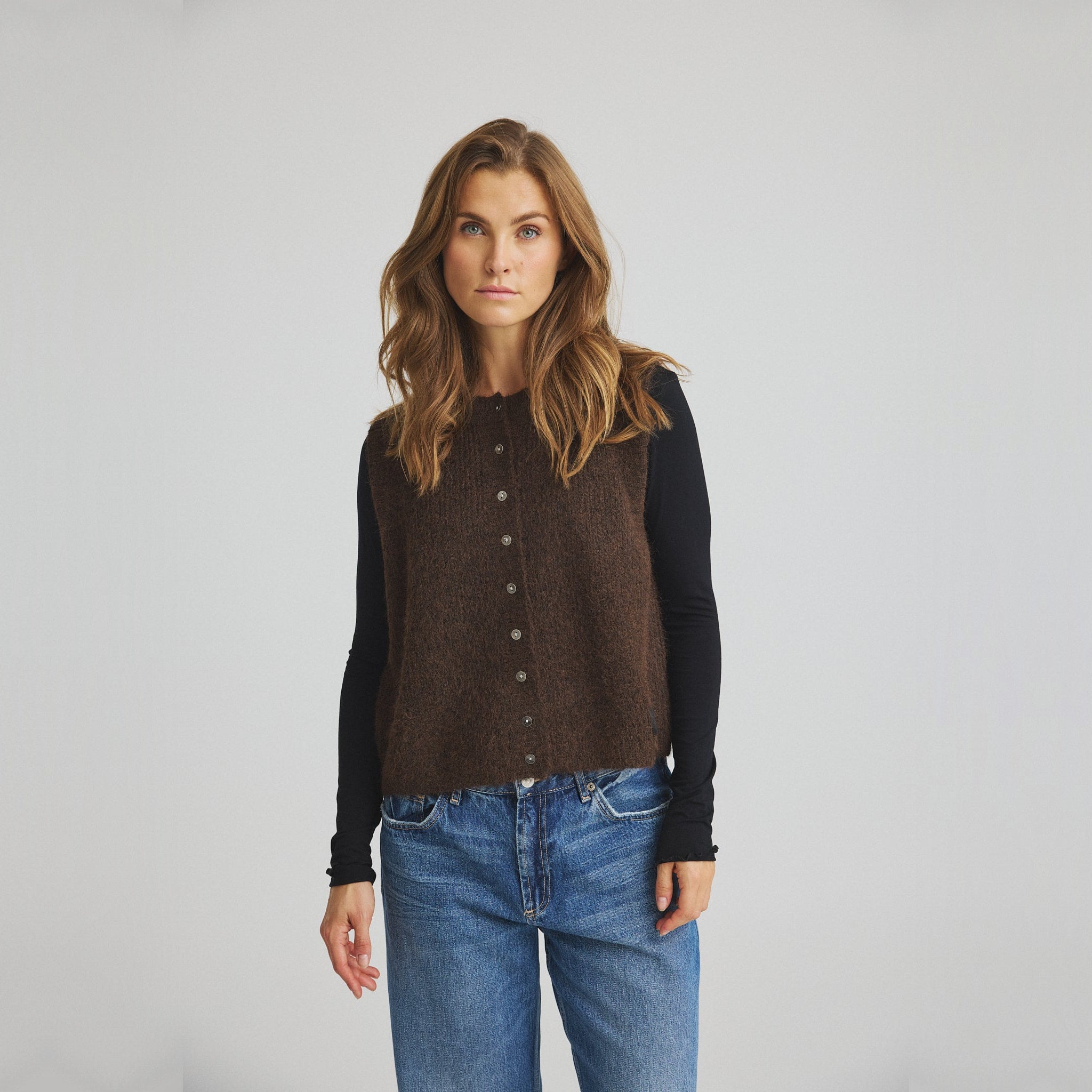 Model i brun strikket vest med knapper over sort langærmet bluse og blå jeans