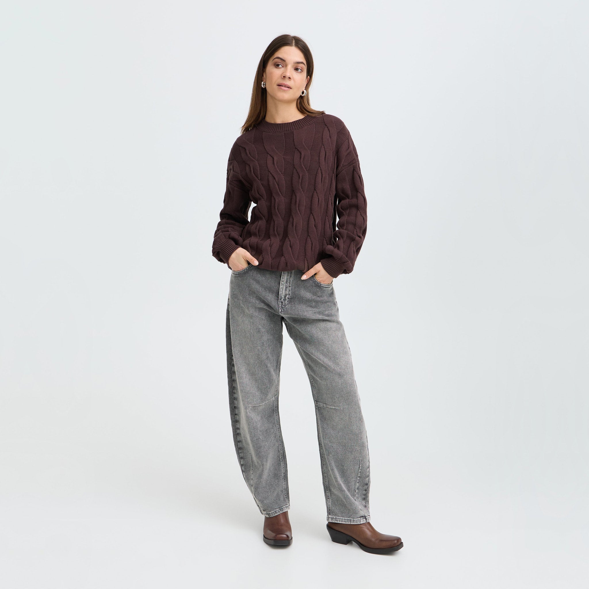 Model i grå barrel leg jeans med brun strikbluse og brune støvler, hele figuren