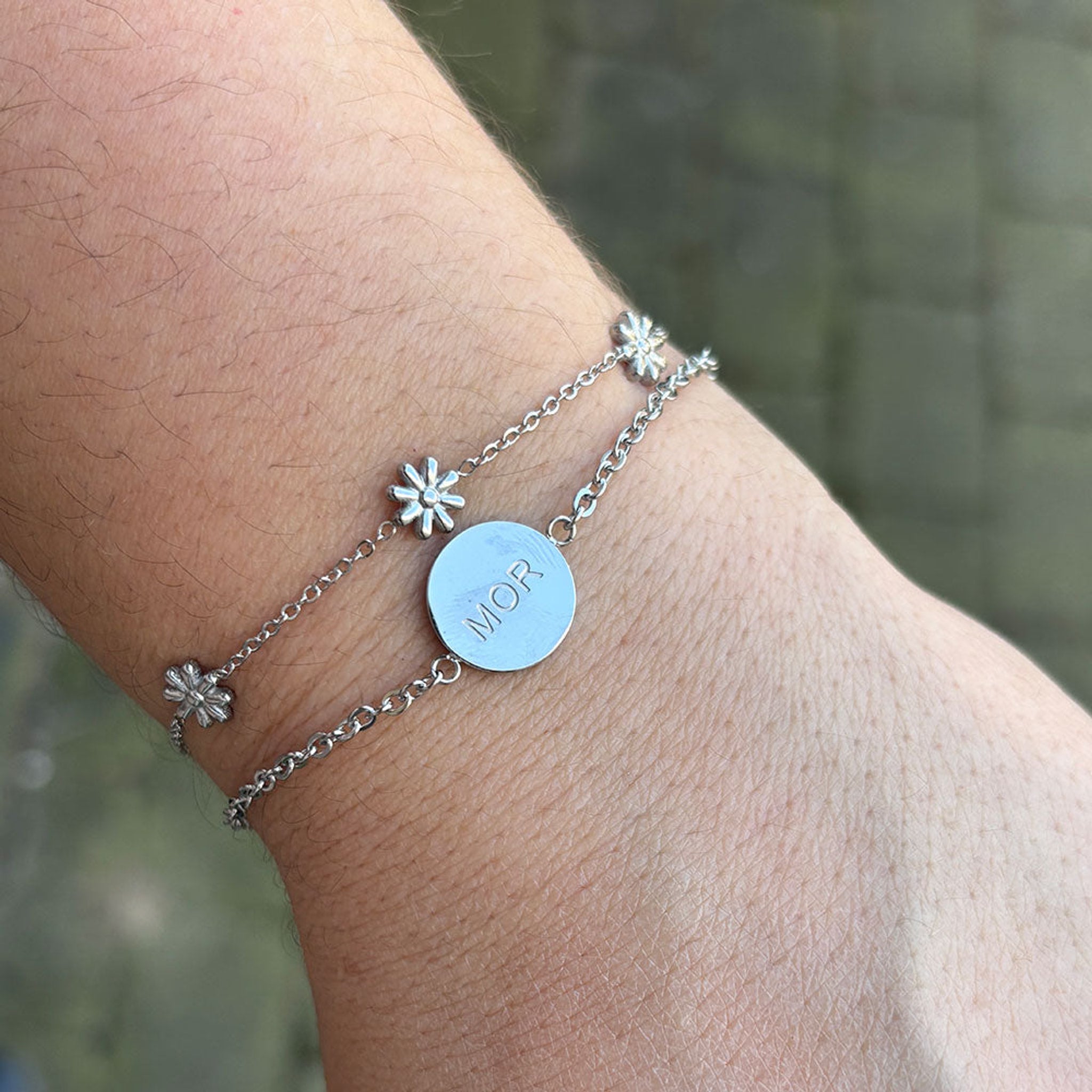 Sølvfarvet armbånd med rund plade med teksten MOR på håndled, stylet med et sølvfarvet kædearmbånd med blomster