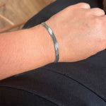 Sølvfarvet armbånd med flad kæde båret på håndleddet mod sorte bukser