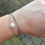 Sølvfarvet armbånd med hvid perle på håndled, stylet sammen med et snoet kædearmbånd