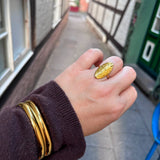 Mindy Bangle armbånd i guld båret om håndleddet oven på ærmet af en brun strikbluse