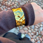 Bredt armbånd i guld med bulet overflade båret uden på ærmet på en brun strik