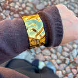 Bredt armbånd i guld med bulet overflade båret uden på ærmet på en brun strik