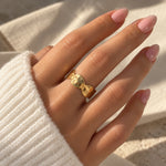 Bred ring i guld med hamret overflade båret på en finger tæt på, sammen med en hvid strikbluse
