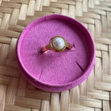 Guldfarvet Sally Mini Pearl Ring med lille hvid perle i rund indfatning, placeret i en pink ringholder