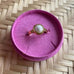 Guldfarvet Sally Mini Pearl Ring med lille hvid perle i rund indfatning, placeret i en pink ringholder