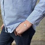 Sølvfarvet armbånd med rund mønt på håndleddet, stylet med blåstribet skjorte og mørke jeans