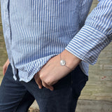Sølvfarvet armbånd med rund mønt på håndleddet, stylet med blåstribet skjorte og mørke jeans