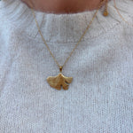 Halskæde i guld med ginkgoblad-vedhæng båret oven på en lys strik, tæt på