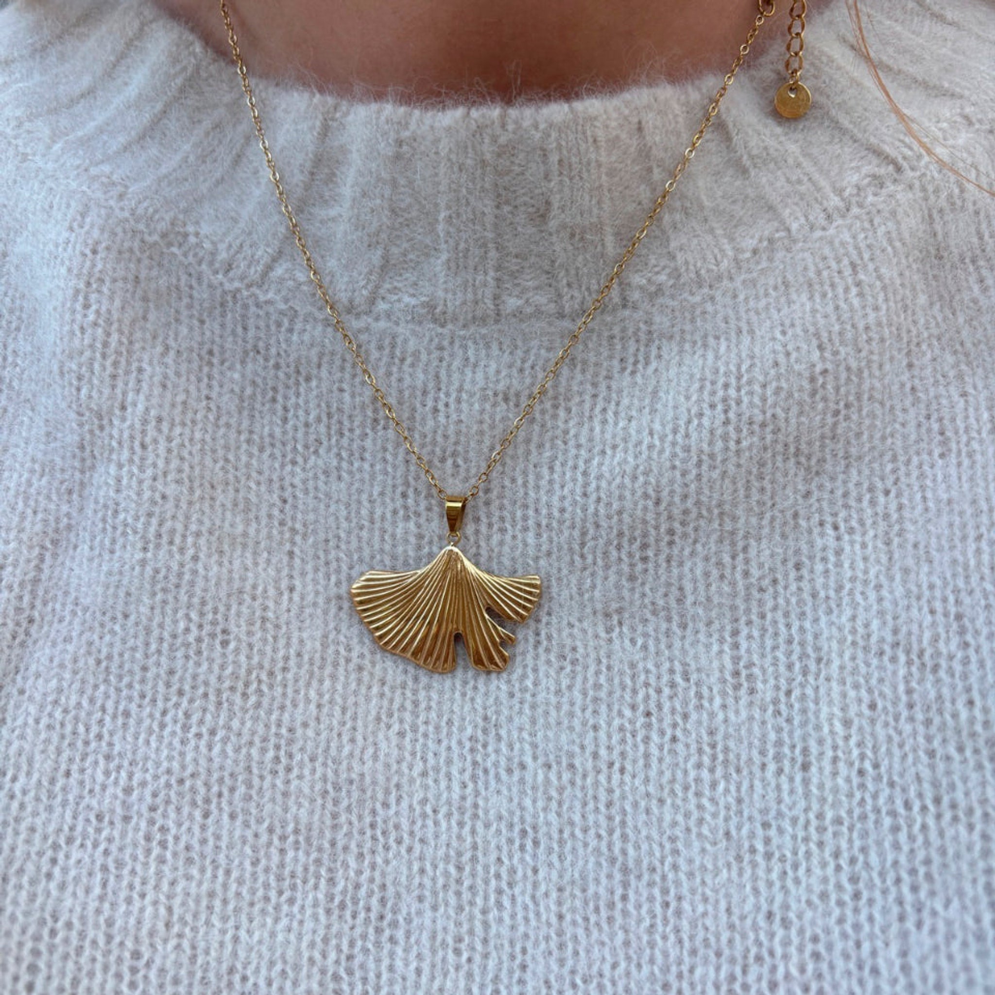Halskæde i guld med ginkgoblad-vedhæng båret oven på en lys strik, tæt på