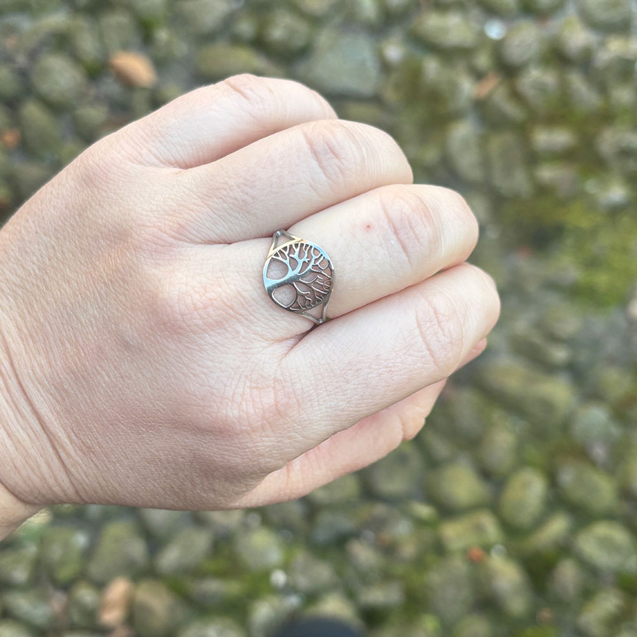 Sølvfarvet ring med livets træ båret på en finger, set tæt på mod en stenmur
