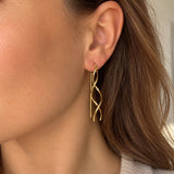 Model bærer Waves ørering i guld, hvor de to bølgede stave snor sig som en spiral under øreflippen, set fra siden